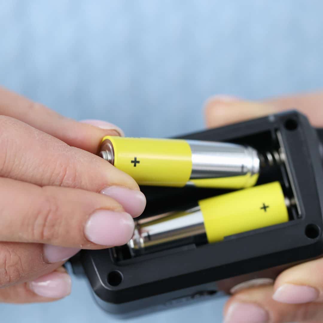 A hand inserting a yellow-wrapped AA battery into a black electronic device.