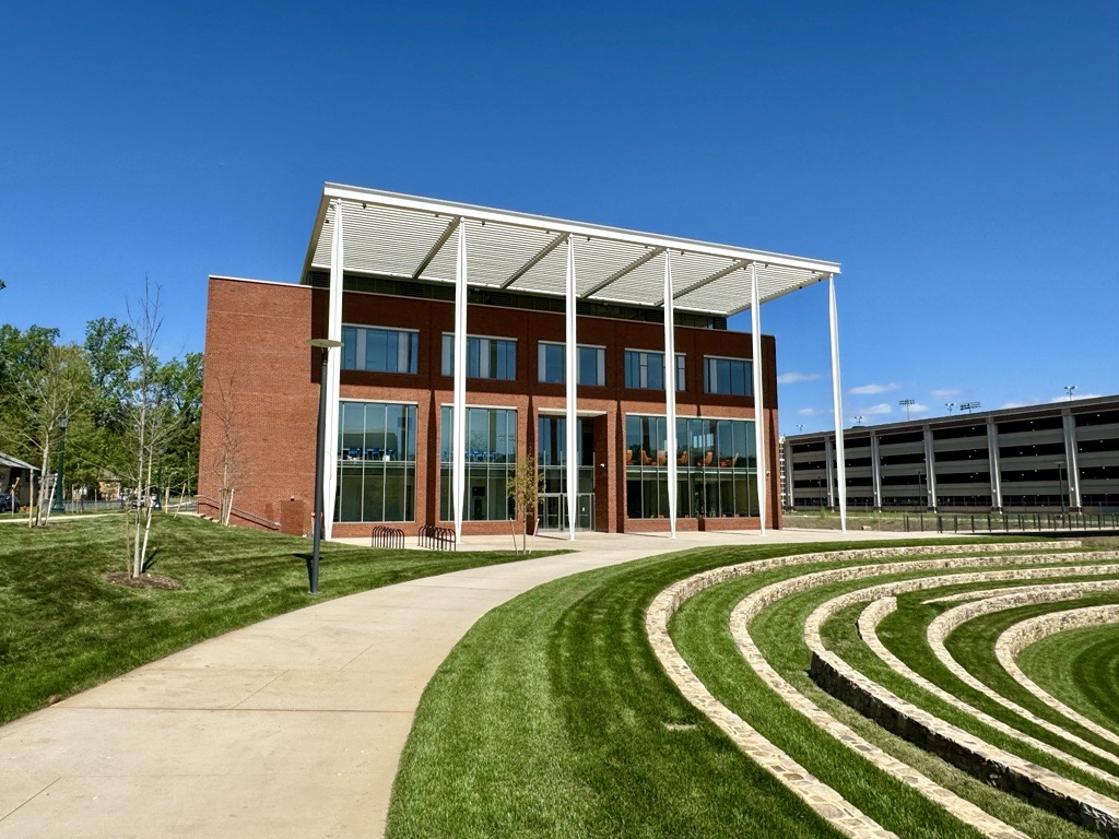 Red brick building with tall white columns and large windows, set against a clear blue sky. Curved grassy steps are in the foreground.