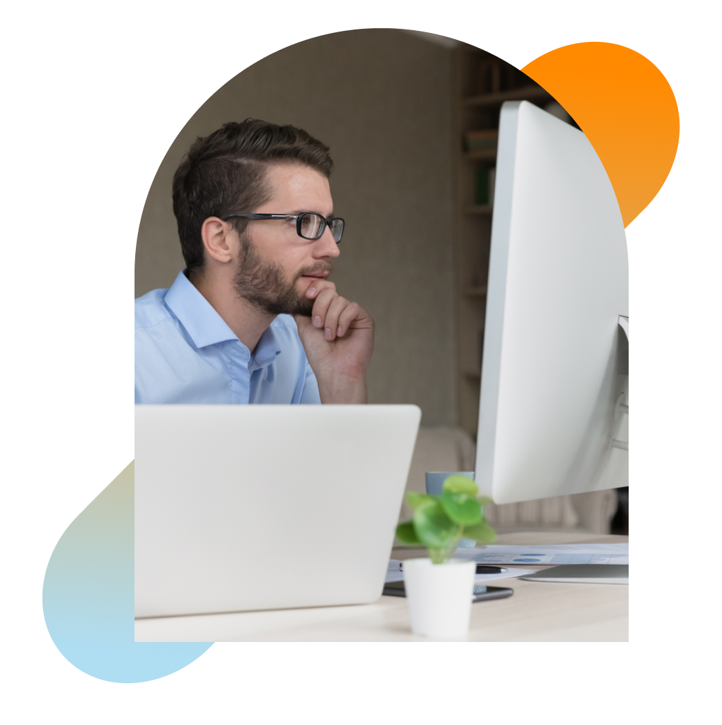 Contemplative man wearing glasses seated in front of computer, staring at large monitor.