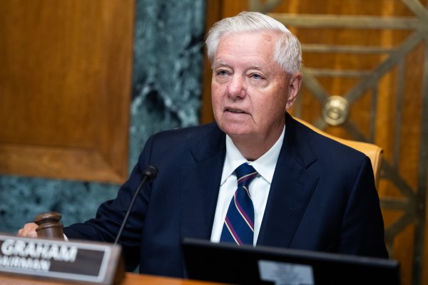 An elderly man in a dark suit and striped tie speaks at a microphone during what appears to be a formal legislative or governmental hearing in an ornate chamber.
