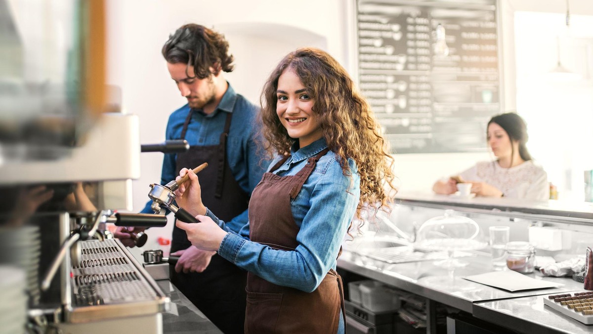 Studenten bei der Arbeit. Die Minijobregelung soll künftig nur noch für Schülerinnen und Schüler gelten. Dagegen gibt es Kritik. Two young baristas working in coffee shop, standing by counter. University students working part-time in cafe.