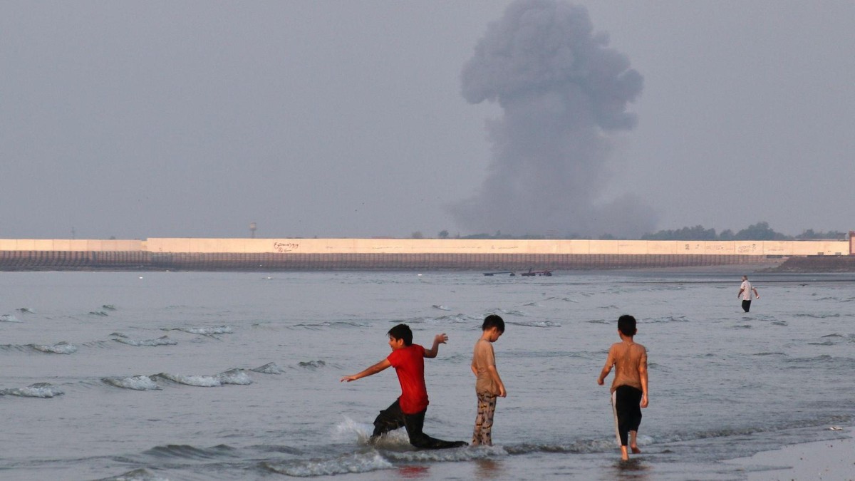 Drei Jungen spielen im seichten Wasser der Straße von Hormus, während im Hintergrund vor der Küste von Bandar Abbas im Iran eine Rauchwolke nach einer Explosion aufsteigt. (zu dpa: «Bericht: Haus von Wildhüter im Iran bombardiert») Iran-Krieg - Straße von Hormus
