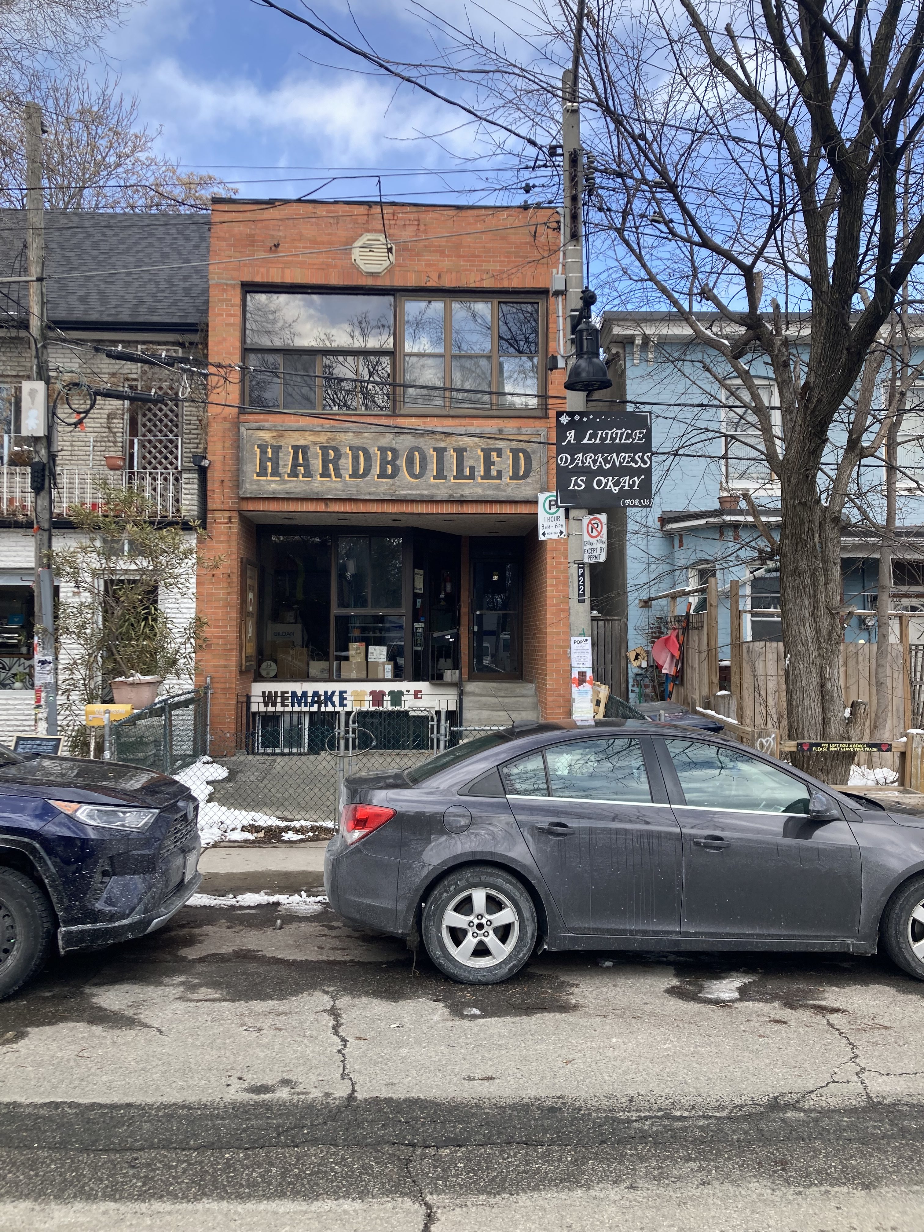 An old brick two-storey shop in Toronto's Kensington Market neighbourhood fronts a wide sign with block lettering that reads "HARDBOILED," while a smaller, square sign in front of it affixed to a utility pole reads "A LITTLE DARKNESS IS OKAY" in a stylized script font. Blue and gray cars are parked at the curb in front of the building.