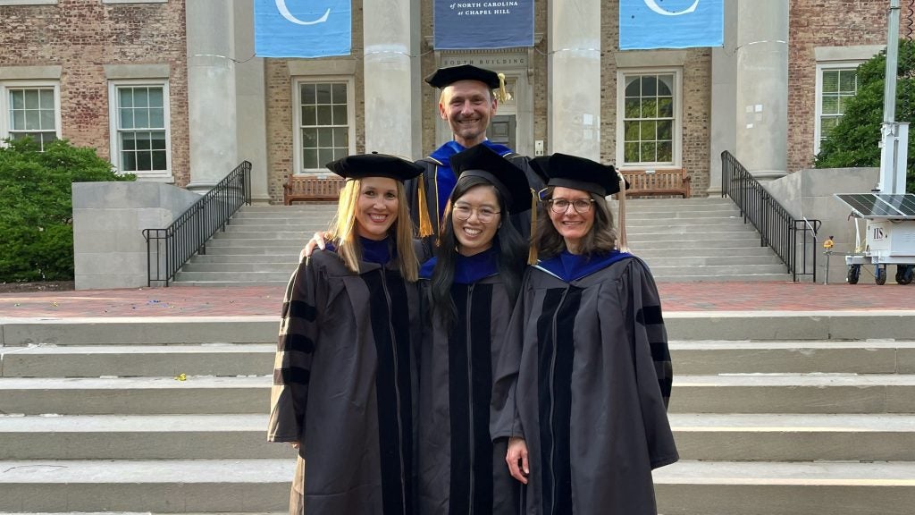 Photo of three PhD graduates with their advisor in their regalia outside South Building on UNC campus.