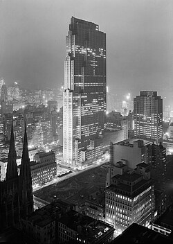 A monochrome image of a building being constructed, with lit office rooms especially prominent