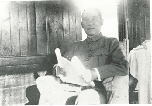 A black and white photograph of a seated man wearing a military or pseudo-military jacket