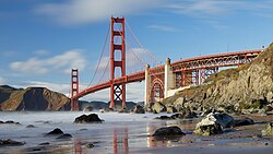 The Golden Gate Bridge, a two-tower suspension bridge painted a dull orange, seen on a sunny day from a beach with mist over the water.