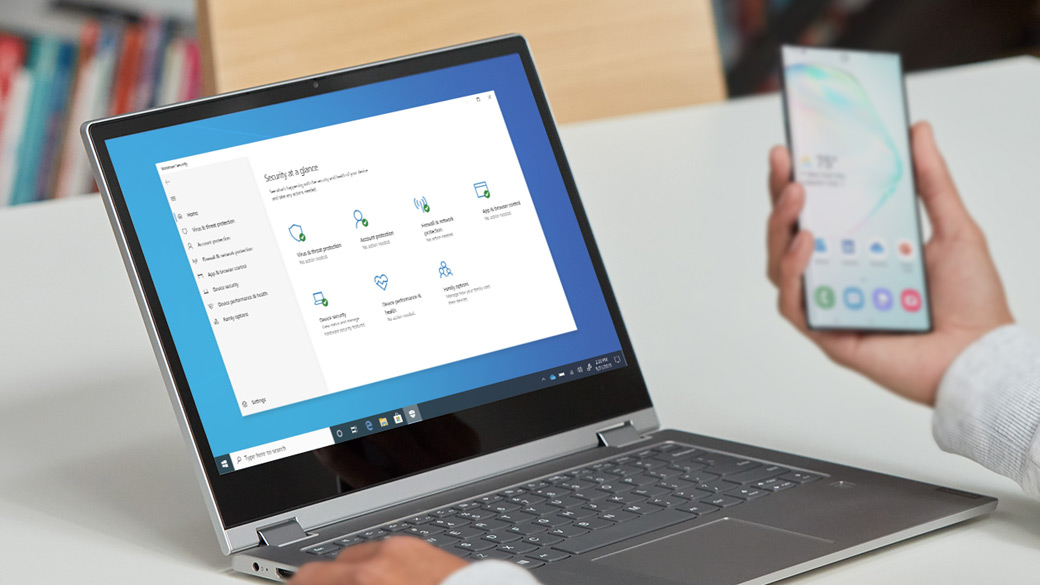 Person reviews cell phone while Windows 10 laptop displays security features