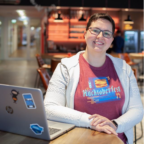 Woman wearing a Hacktoberfest T-shirt resting her arm on a desk next to her open laptop.