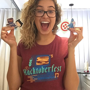 Woman wearing Hacktoberfest T-shirt holding up Hacktoberfest stickers with a big open mouth smile.
