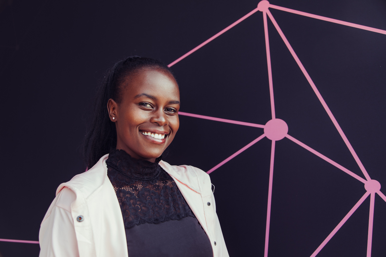 Photo of Sonia John smiling in front of a GitHub Universe backdrop. The pink graphics matches their pink jacket.