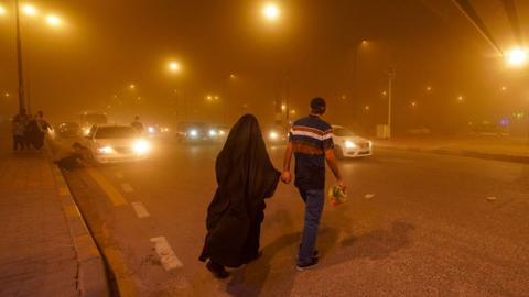 During a sandstorm in Najaf, Iraq on 14 April 2024, a man dressed in a black shirt with red and white stripes walks hand in hand with a woman dressed in a black burka. They are crossing a busy road with cars as thick, orange cloud blankets the air and lights from street lamps shimmer around them.