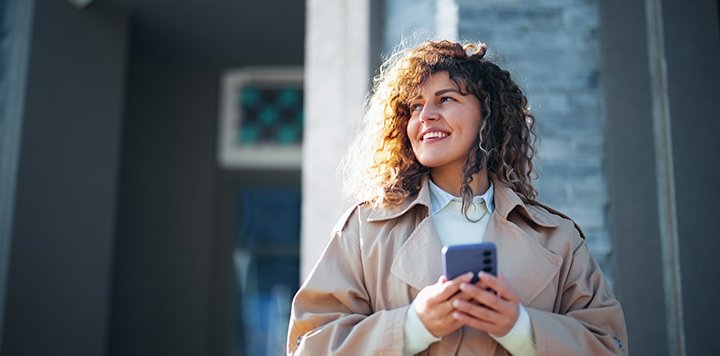 A young woman with curly hair, wearing a beige trench coat, smiles whilst holding a mobile phone outdoors. She looks off to the side, with sunlight highlighting her face. The background features a grey building with a modern design.