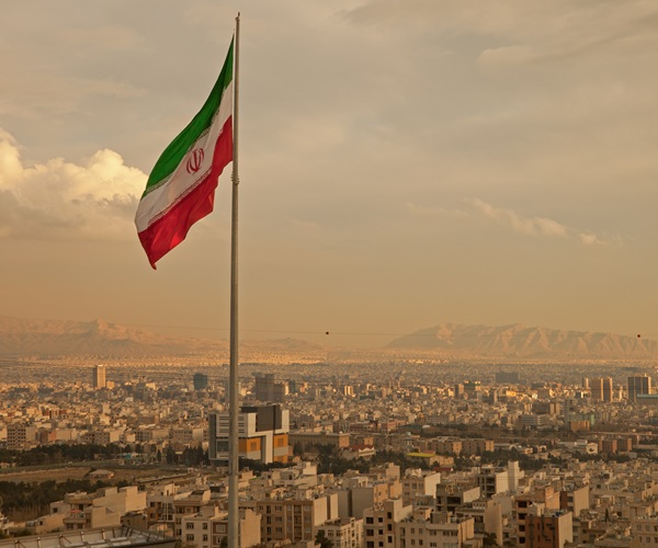 Iran flag waving in the wind above skyline of Tehran lit by orange glow of sunset.