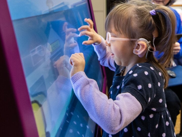 A little girl wearing pink glasses is placing both of her hands on a large screen.
