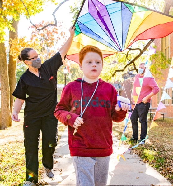 A visually impaired boy with bright red hair flying a colorful kite outside.