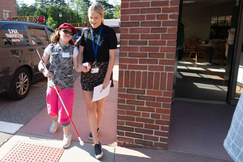 A Perkins' volunteer walking with a visually impaired girl with a guide cane outside.