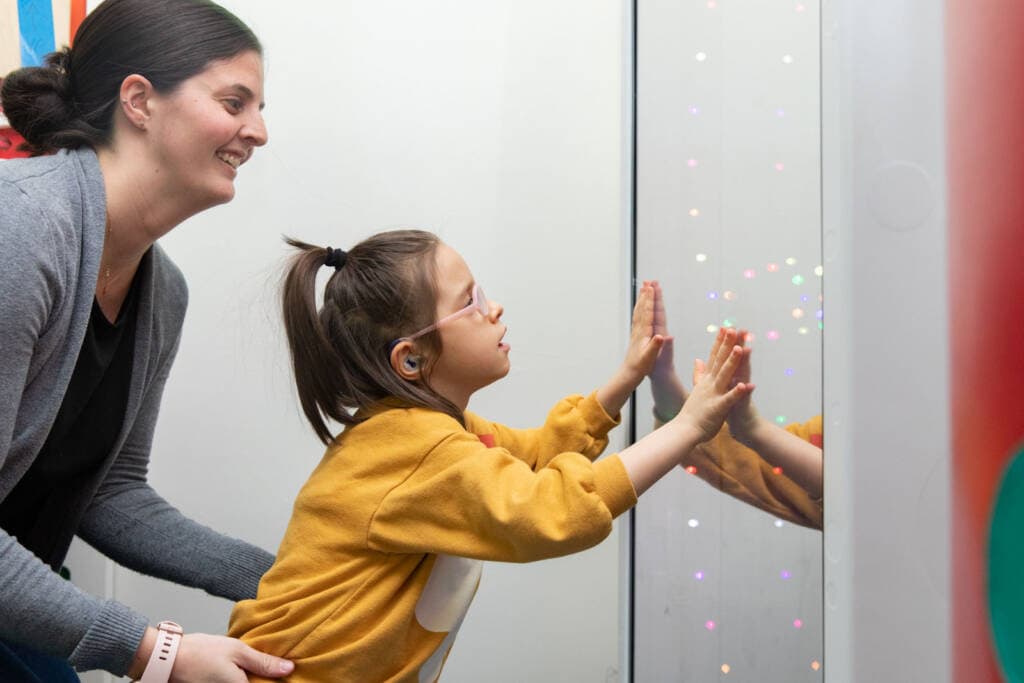 A girl puts her hands on a light display window while her teacher supports her from behind.