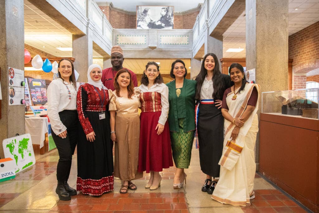 Group photo of international scholars containing 7 women and 1 man. Most are dressed in traditional clothes from their home country and pose indoors in an open hallway.