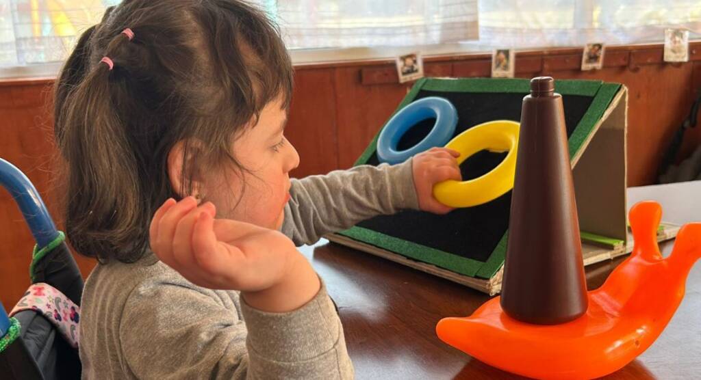 A young child sits at a table, focused on a game involving colorful rings and a cone. The child holds a yellow ring, poised to place it on the brown cone, which is mounted on an orange, snail-shaped base. The setting appears to be indoors.