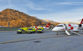 Zwei Rega-Jets und drei Ambulanzfahrzeuge auf dem Rollfeld des Flugplatzes Sion vor einer Bergkulisse.