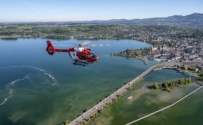 Ein roter Rega-Rettungshelikopter fliegt über einen See und eine Uferstadt in der Schweiz. Im Vordergrund sind Brücken und flaches Wasser zu sehen, während sich im Hintergrund eine hügelige Landschaft unter klarem Himmel erstreckt.